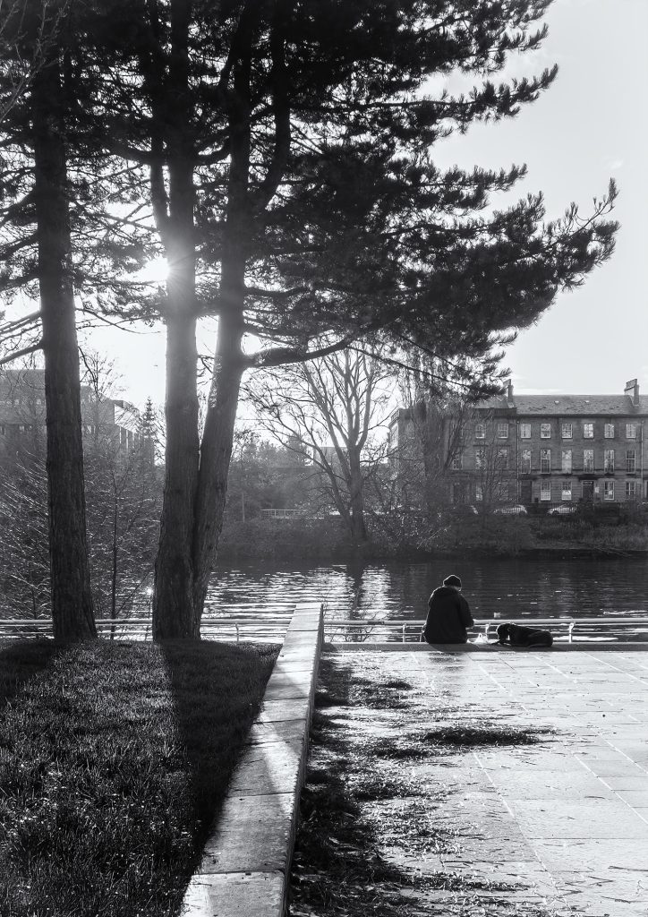 A black and white photograph of a person sitting with a dog beside a river with their back to the camera. There are trees to the left of the person and the sun is shining through the trees.