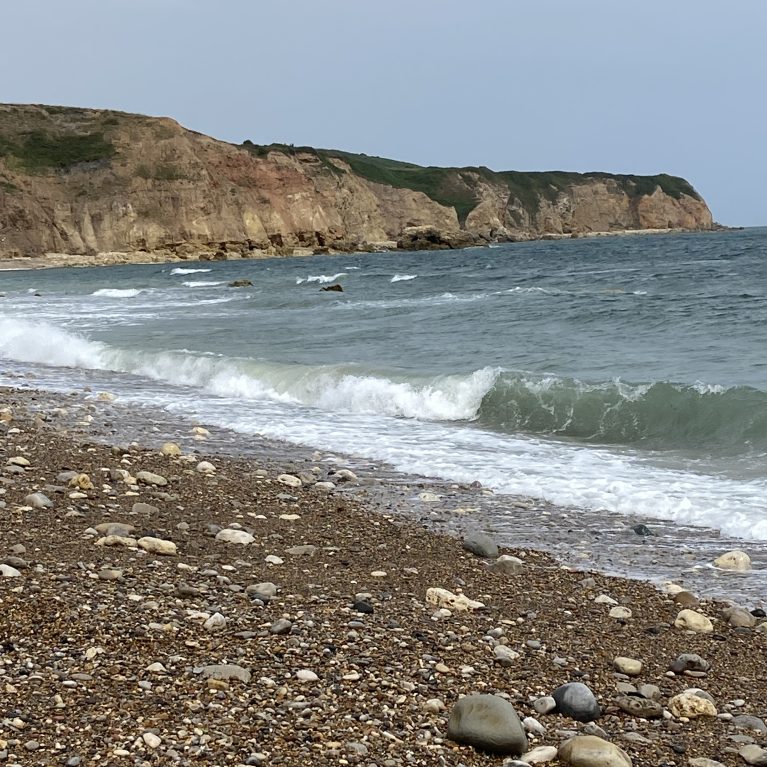 A photograph of a beach with rocks and pebbles, the sea and a cliff in the background.