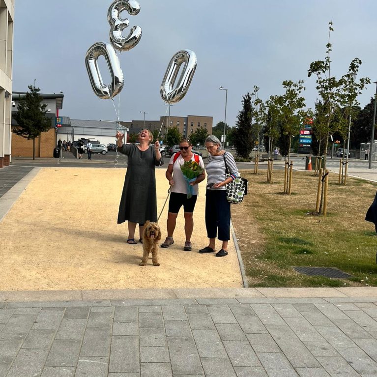 Three people standing in the sun holding balloons with the numbers three, zero and zero