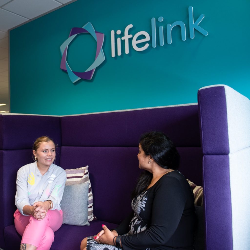 Two people sitting on a sofa under a sign for LifeLink