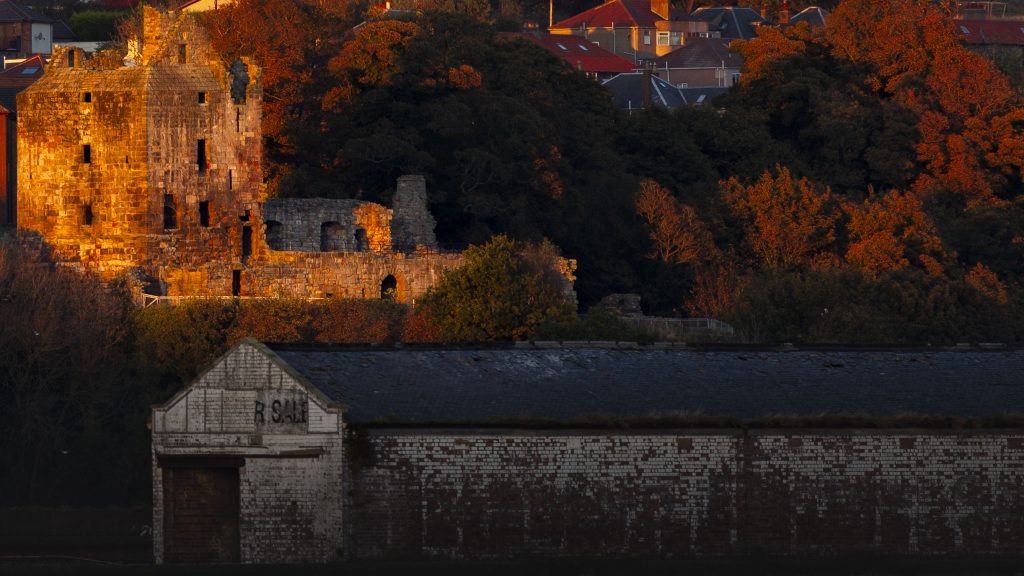 Ravenscraig Castle is lit up by the sun in the left side of the image. The right side is filled with trees in shadow.