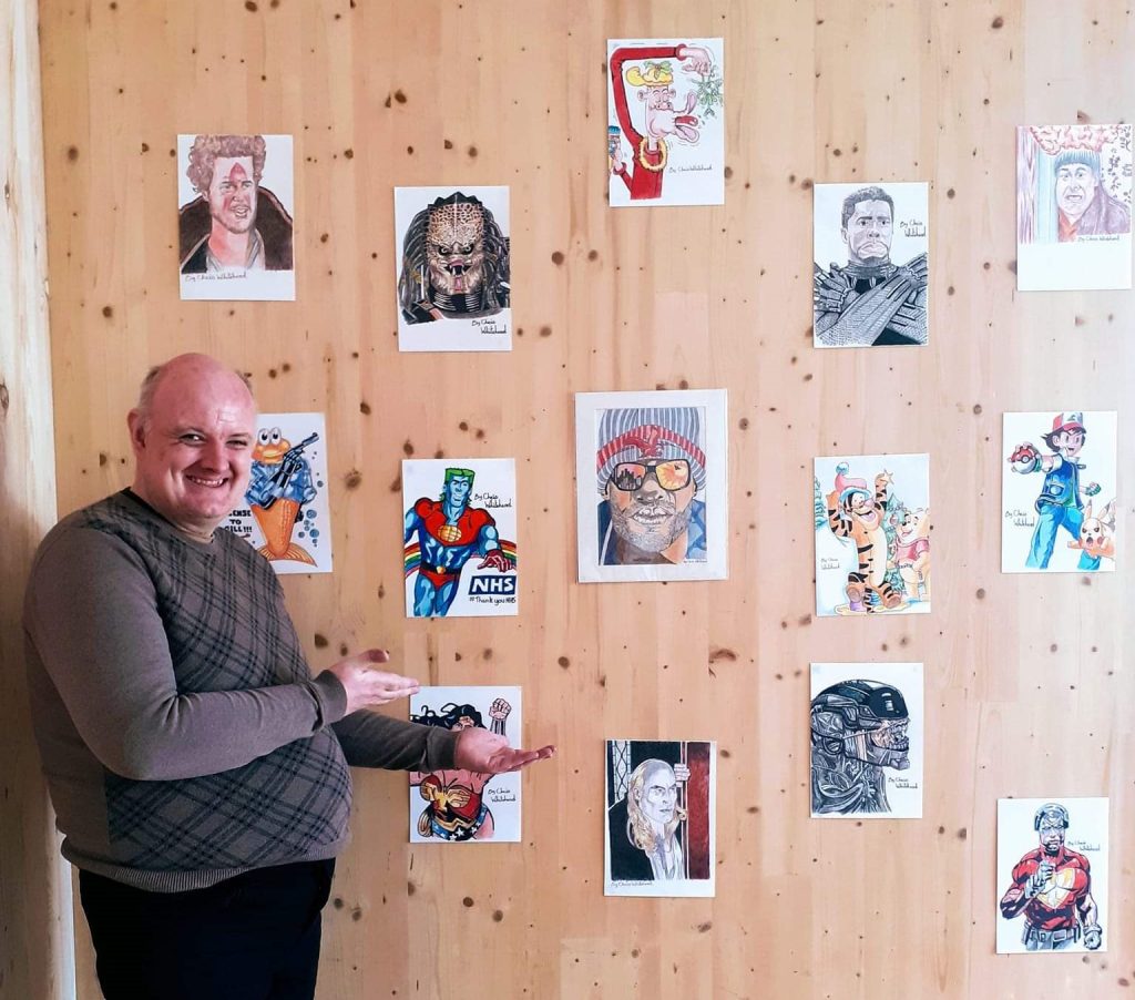 A man smiling at the viewer. He is pointing to pictures on a wall