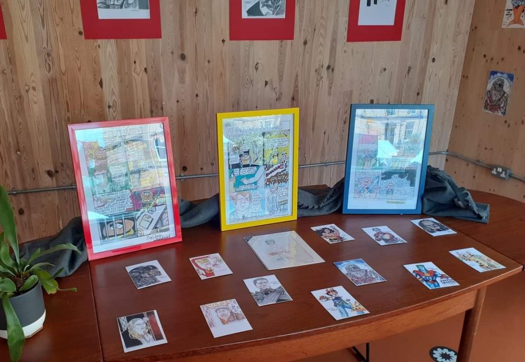 A table with postcards laid out on it and three framed comic pages standing upright on the table.