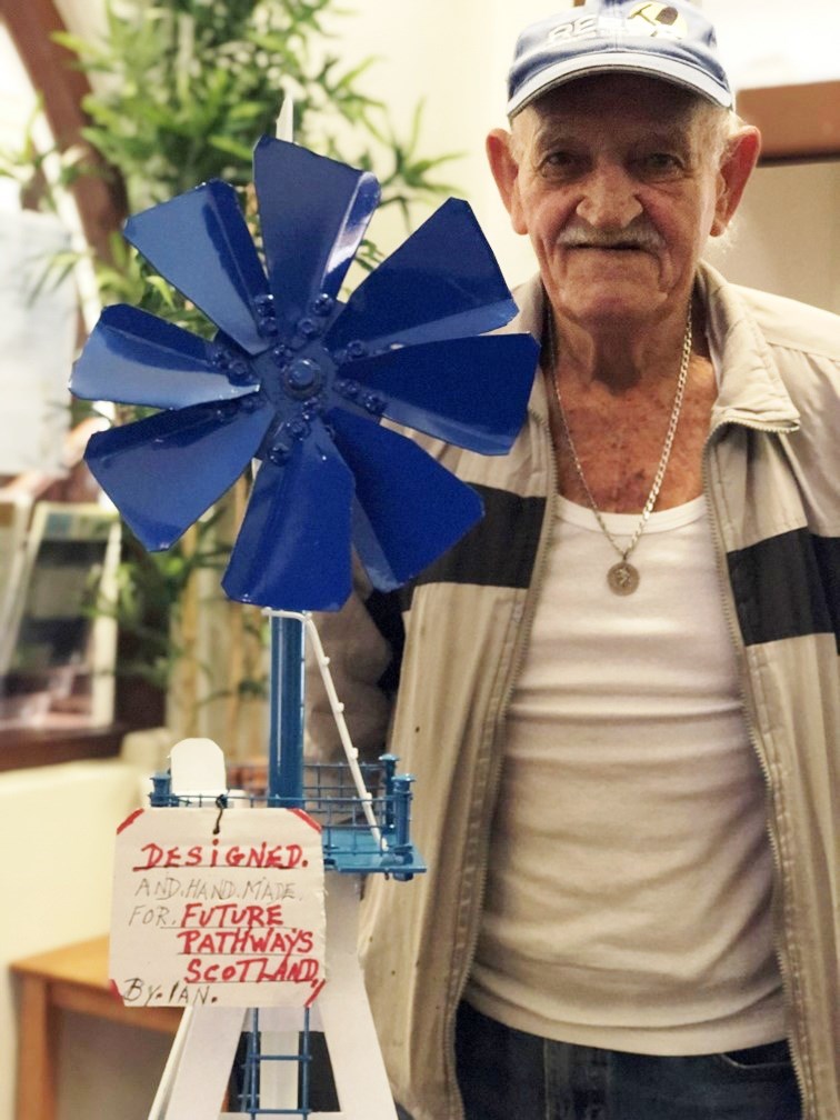 A man wearing a hat, jacket and t shirt looks directly at the viewer. He is holding a handmade windmill which is white with blue sails.