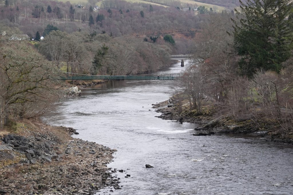 A river running through the countryside with banks of pebbles either side and trees.