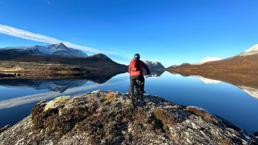 A man on a bike facing away from the viewer. He is looking out over a loch and mountains.