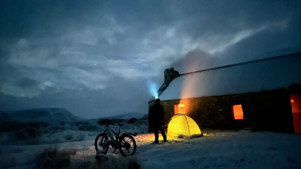 A man stands with a bike and tent at night time at Loch Vaich.