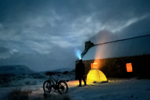 A man stands with a bike and tent at night time at Loch Vaich.