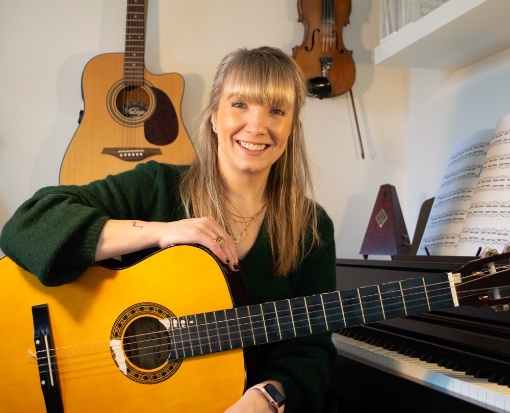 A woman holding a guitar and smiling into the camera.