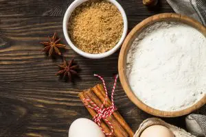 A bowl of sugar, a bowl of flour, cinamon sticks and eggs on a table.